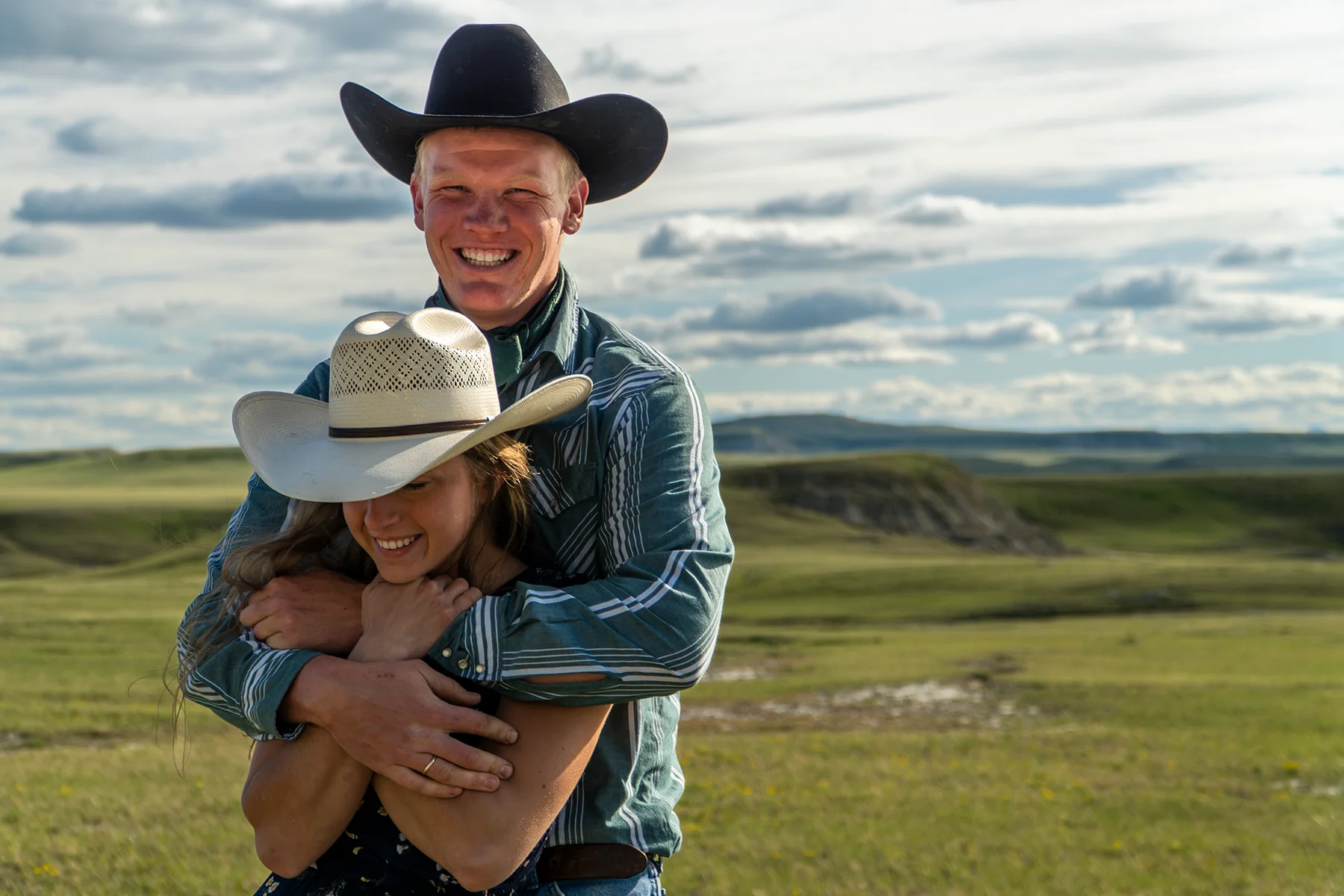 A smiling couple in cowboy hats embrace in an open grassy landscape